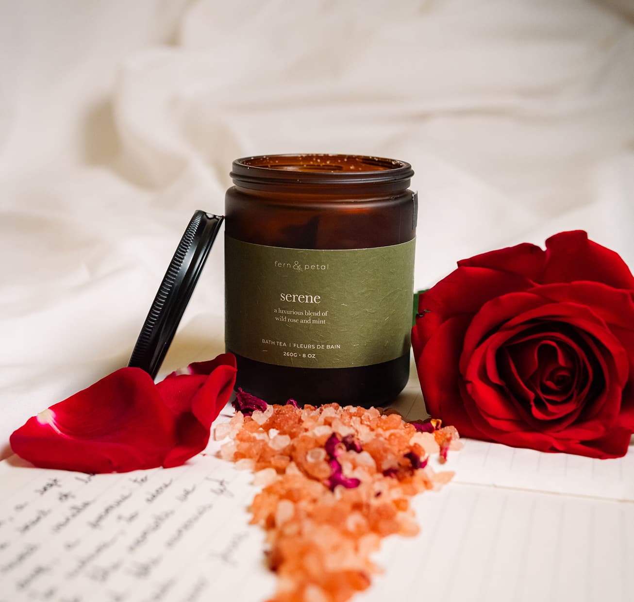 An open container of serene with bath salts in front of it and a rose flower beside it.